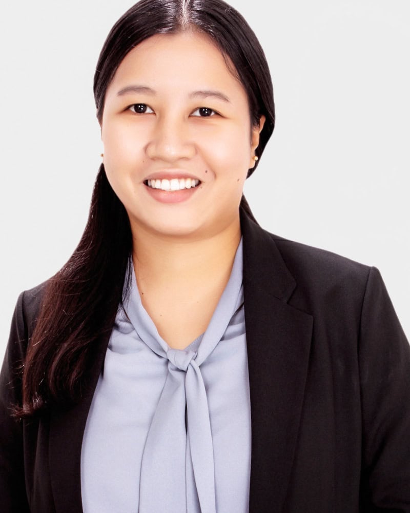 A woman with long dark hair, wearing a black blazer over a light blue blouse, smiles at the camera against a plain white background.