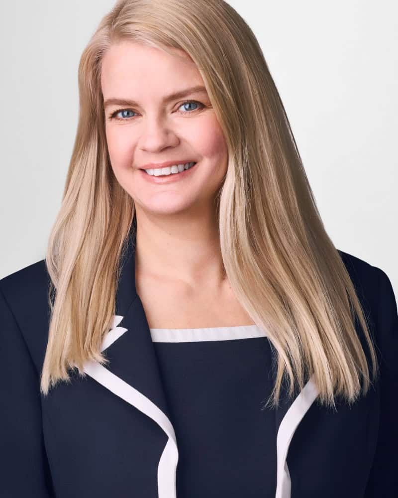 A woman with long blonde hair, wearing a dark blazer with white trim, smiles at the camera against a plain light background.