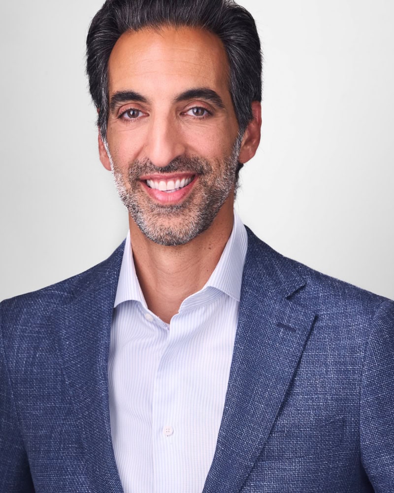 A man with dark hair and gray streaks, a short beard, and a friendly smile, wearing a white shirt and a blue textured blazer, poses against a plain light background.