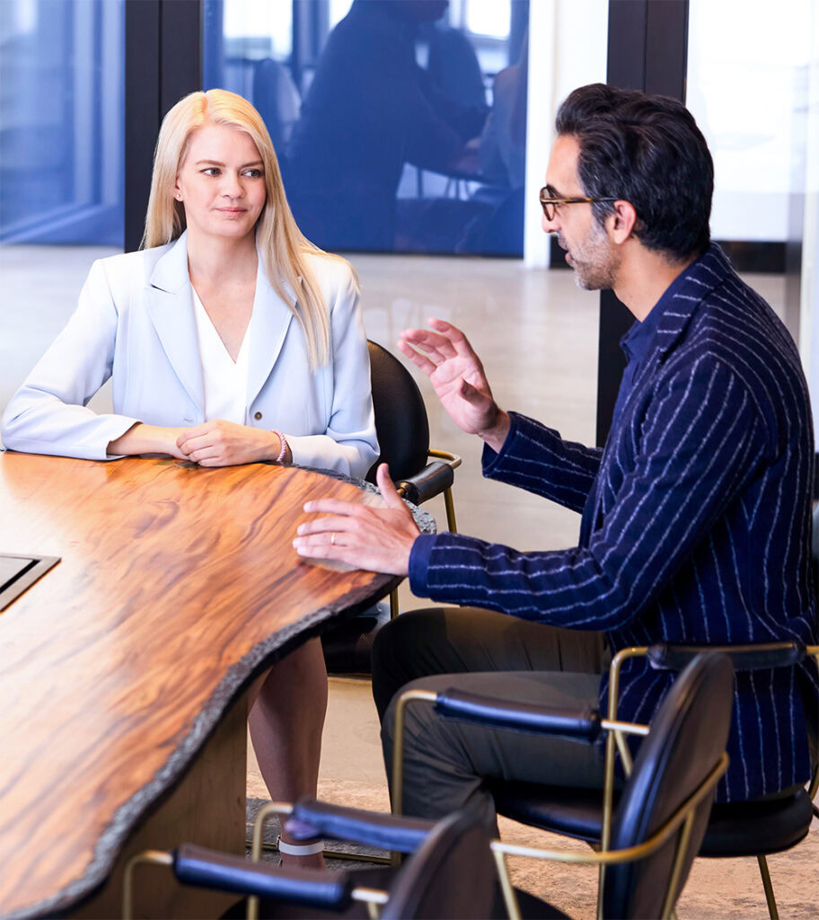 A woman in a light blazer and a man in glasses and a dark striped jacket are sitting at a wooden conference table, engaged in conversation in a modern office setting.