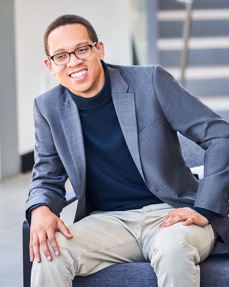 A young man wearing glasses, a gray blazer, a dark turtleneck, and light-colored pants sits smiling on a chair indoors, with a staircase and bright background behind him.