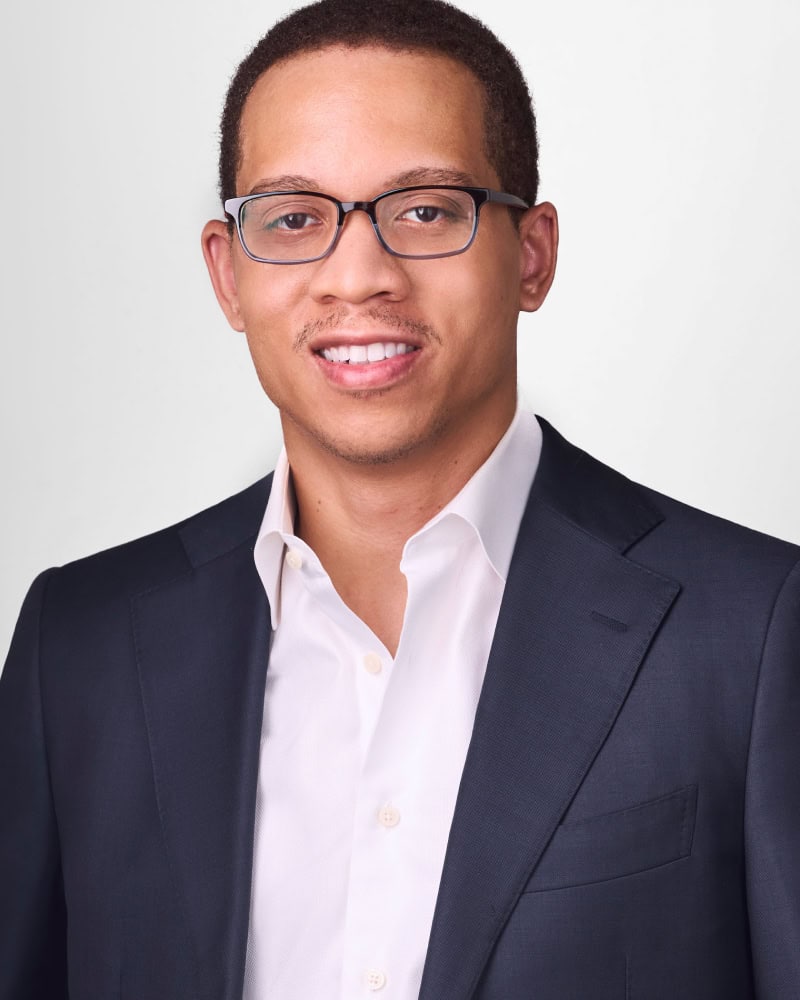 A man wearing glasses, a dark suit jacket, and a white dress shirt, smiling at the camera against a plain light background.