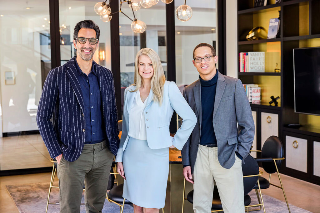 Three professionally dressed people stand smiling in a modern office with glass walls, a round table, chairs, built-in shelves, and decorative lighting in the background.